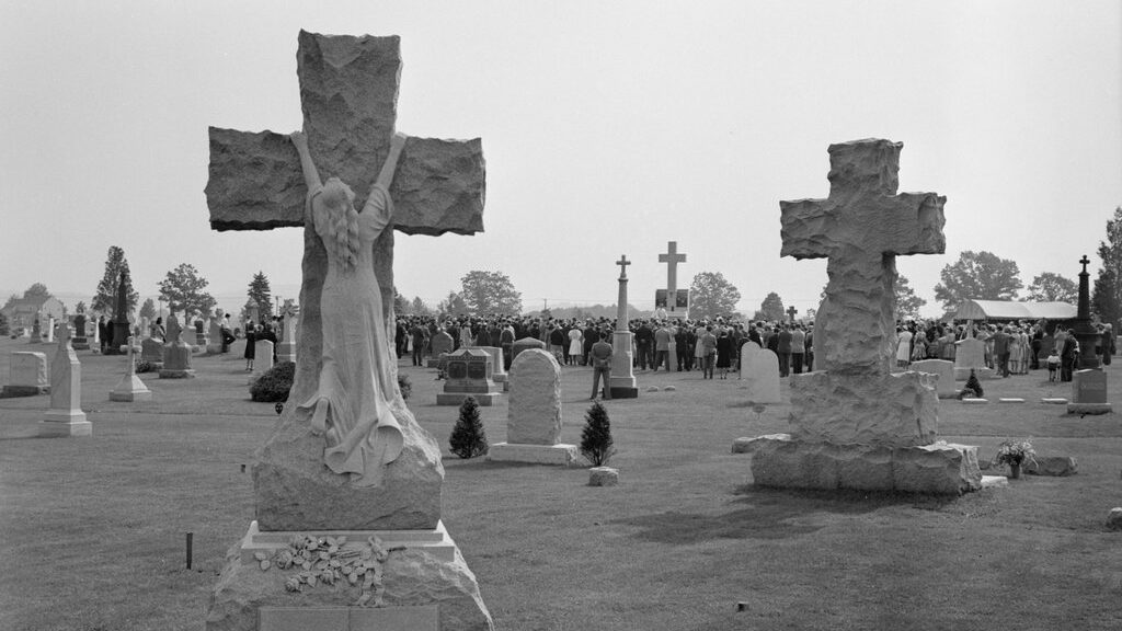 All Souls' Day Mass at Saint Thomas Cemetery in Southington, Connecticut. A crowd of people is gathered near a large cross in the background. In the foreground, large carved crosses and a prominent statue of a woman kneeling against a cross.