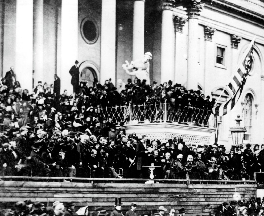 President Abraham Lincoln, surrounded by hundreds of supporters and government officials, delivers his second inaugural address on the Eastern portico of the U.S. Capitol. A large American flag is prominent on the balcony just to his left.