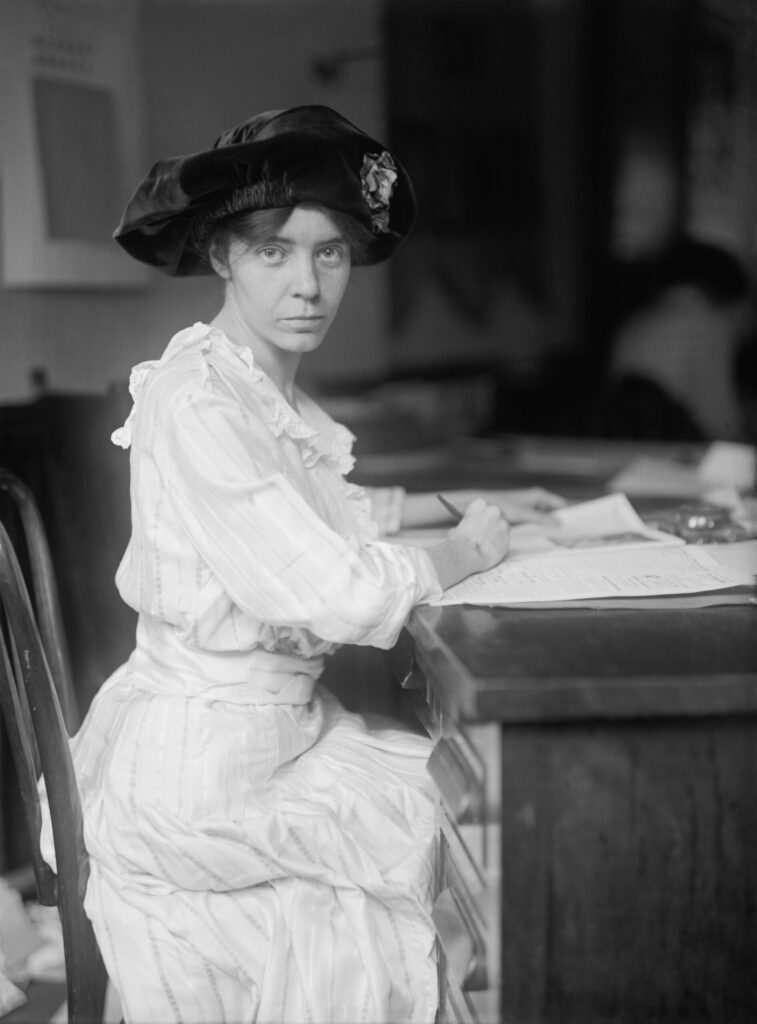 Photograph of Quaker and suffragette Alice Paul, 1915. Paul is shown seated at a desk with pen in hand and paper on a desk. She wears a white dress with a ruffled colar and a black velvet hat. 