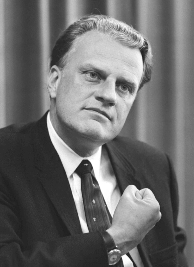 Photograph of Billy Graham seated in a dark suit and tie with cross tie pins and with his right fist clenched to his chest.