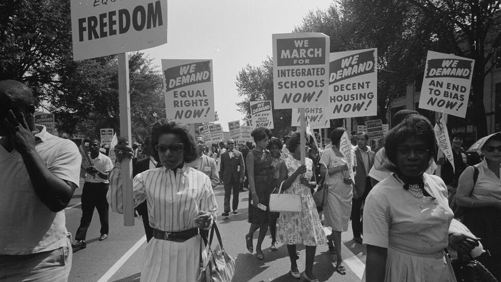 The photograph shows a procession of Black American civil rights marchers with picket signs advocating for better housing, school integration, and equal rights. In the front are sisters Elsie Eatman on the right and Delores Coleman to the left, carrying a sign that is only partially visible but reads "Freedom." Behind these are signs that say, "We Demand Equal Rights Now!; We march for integrated schools Now!; We demand decent housing Now!; and We demand an end to bias now!" Below this last one are the words "Freedom in '63." Other signs appear, but the words are unclear.