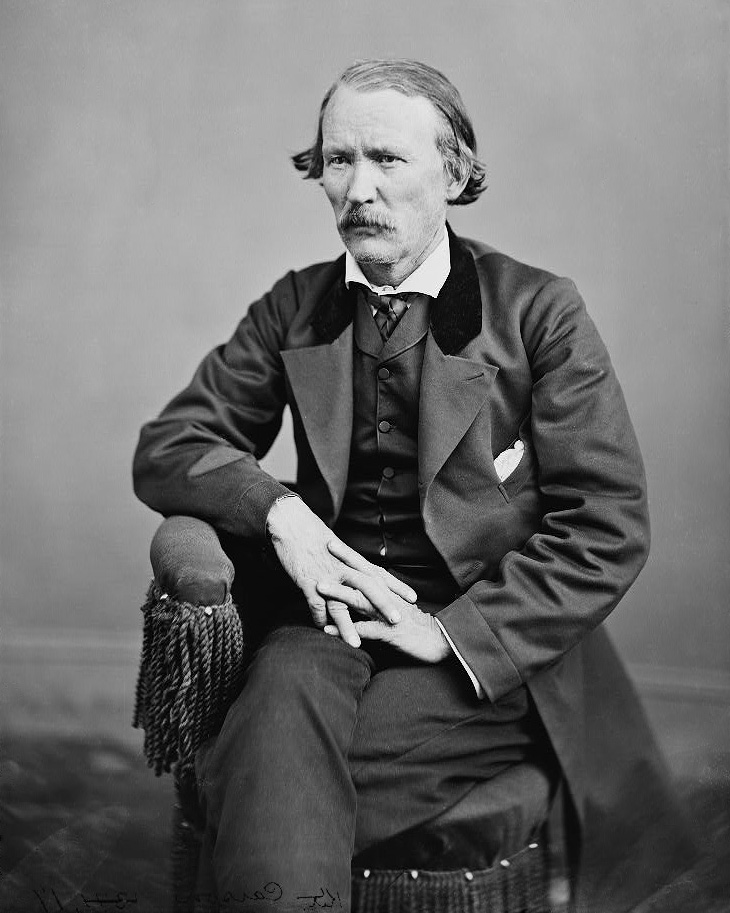 Kit Carson is pictured seated, his hands intertwined in his lap. He is wearing a three-piece suit with a velvet lapel and a white shirt.