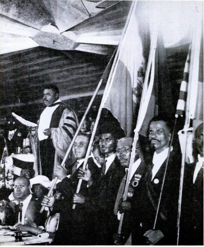 A photograph of Marcus Garvey speaking to an audience in Harlem, NY, 1920. Garvey is wearing academic robes, and to his left are a handful of men holding various flags.