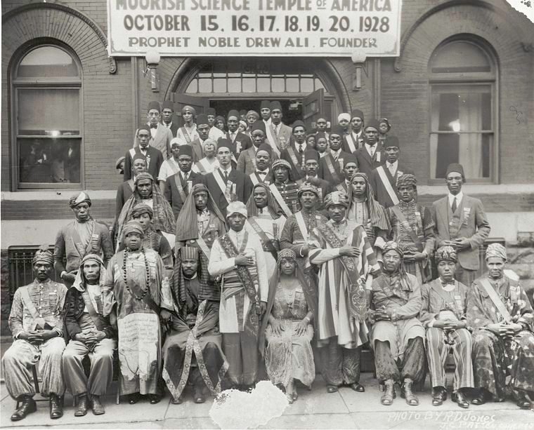 A building with a banner that reads "Moorish Science Temple of America, October 15, 16, 17, 18, 19, 20, 1928. Prophet Noble Drew Ali. Founder." Out front are members of the organization with the Prophet. All members are dressed in clothing from their homeland. Some wear caps and sashes with tunics, while others wear turbans and traditional Egyptian and Saudi headdresses.