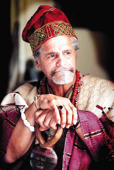 Efuntola Oseijeman Adelabu Adefunmi, the first African American to become part of the Yoruba priesthood, is shown seated in a red cap with a heavily embroidered green border and a tan tunic with colorful green and maroon work. His lands rest on a wooden staff, and he has beads around his neck.