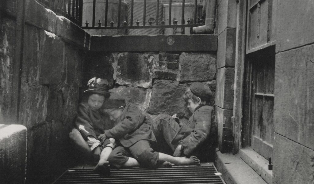 This black-and-white photograph, taken by Jacob Riis, shows three young Arab boys presumed to be homeless sleeping on a grate near a tenement on Mulberry Street, 1890.
