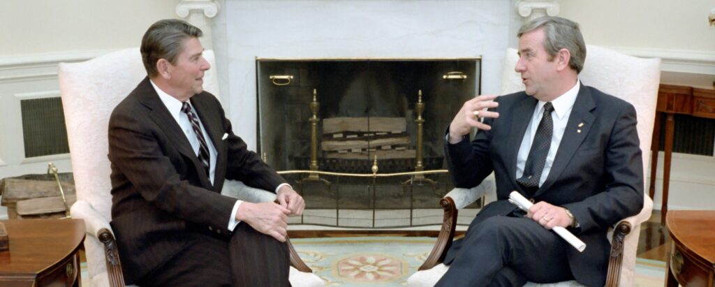 President Ronald Reagan meeting with Baptist pastor and televangelist Jerry Falwell in the Oval Office. Both are seated upon white wingback chairs with carved wood arms. President Reagan is seated to your left with his hands upon his left knee. He appears to be wearing a dark brown or black suit, tie, and white shirt. Falwell is seated to his left in a dark gray suit. He clasps a rolled-up paper in his left hand and is using his left hand expressively while speaking. In the background is a fireplace, bronze andirons.Digital Public Library of America.