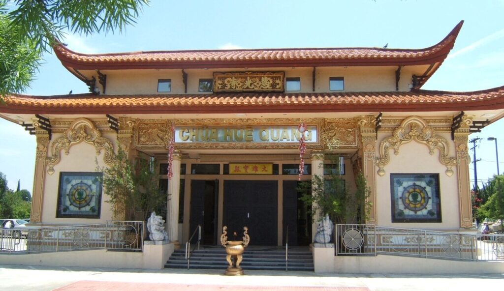 Photograph of a beautiful Vietnamese Buddhist Temple, labeled the "Chua Hue Quang Temple." The temple has two pagodas of a reddish hue. Painted in a soft cream color, the structure is embellished with gold arches on each front section as well as other gold leaf accents and two columns. Two marble lions sit at the main entrance at the top of the stairs. The photograph was taken in 2006 in Garden Grove, California.