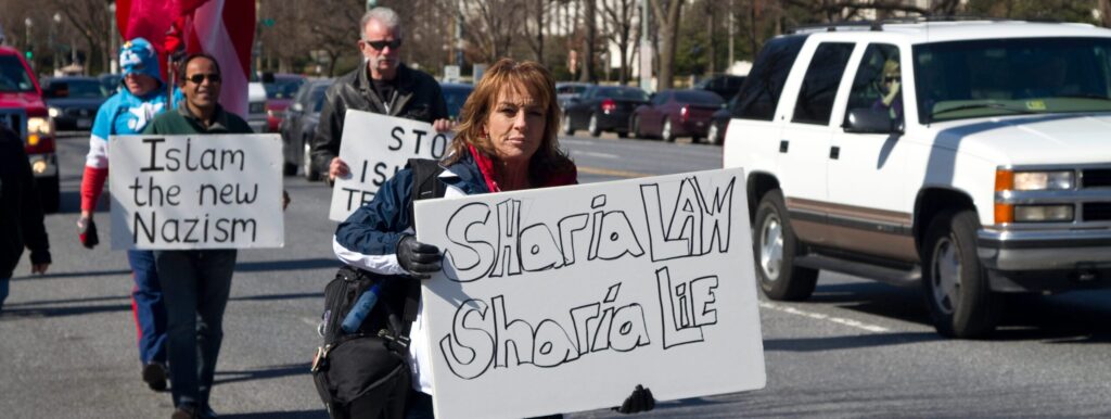 Four Islamophobic marchers in Washington, D.C., including pastor Terry Jones, carry signs vilifying Islamic ideologies. The lady in the front holds a sign that reads "Sharia law, Sharia lie." Behind her is one man carrying a sign that says, "Islam the New Nazism." Pastor Jone's sign is not fully visible put appears to read, "Top Islam Terror." Parked and moving cars are seen in the background.