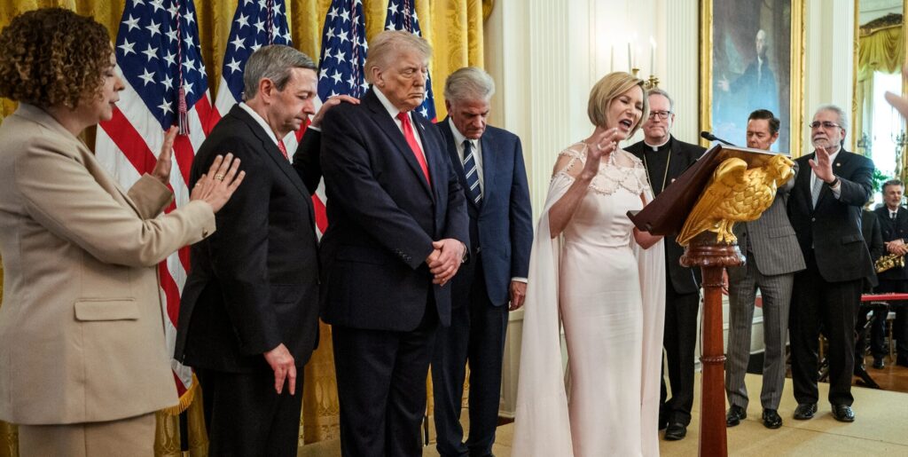 President Trump appears with several conservative religious leaders, including pastor Paula White, who are seen here publicly praying for him. White is in the left center at a pulpit, praying. President Trump is to her right eyes closed and hands clasped.