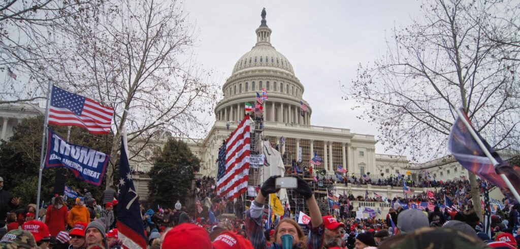 A photograph of the protesters who stormed the US Capitol on January 6, 2021. The white house is in the background. Dozens of protesters hold U.S. Flags and "Trump keep America Great" flags. 