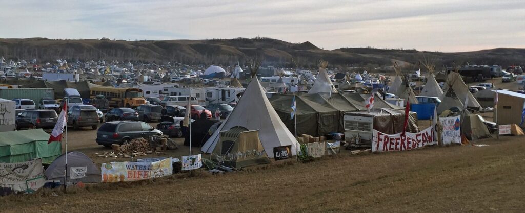 At the Oceti Sakowin camp, people protest the Dakota Access Pipeline at Standing Rock Indian Reservation. Teepees, trailers, buses, and cars are present with flags of various nations. A fence runs in front of the encampment. One sign attached to the fence reads "Water is Life!" and another "Free Red Fawn."