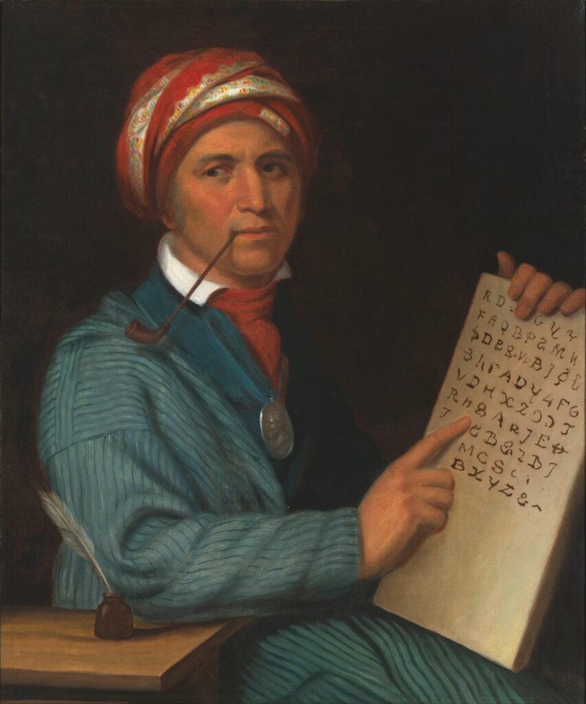 Cherokee Sequoyah is shown with his syllabary. He is wearing a turquoise tunic with lines, a white shirt, a reddish cravat, a silver pendant, and a traditional multicolored head covering. Sequouyah has a slim pipe in his mouth. He is seated at a small table with ink and quill.