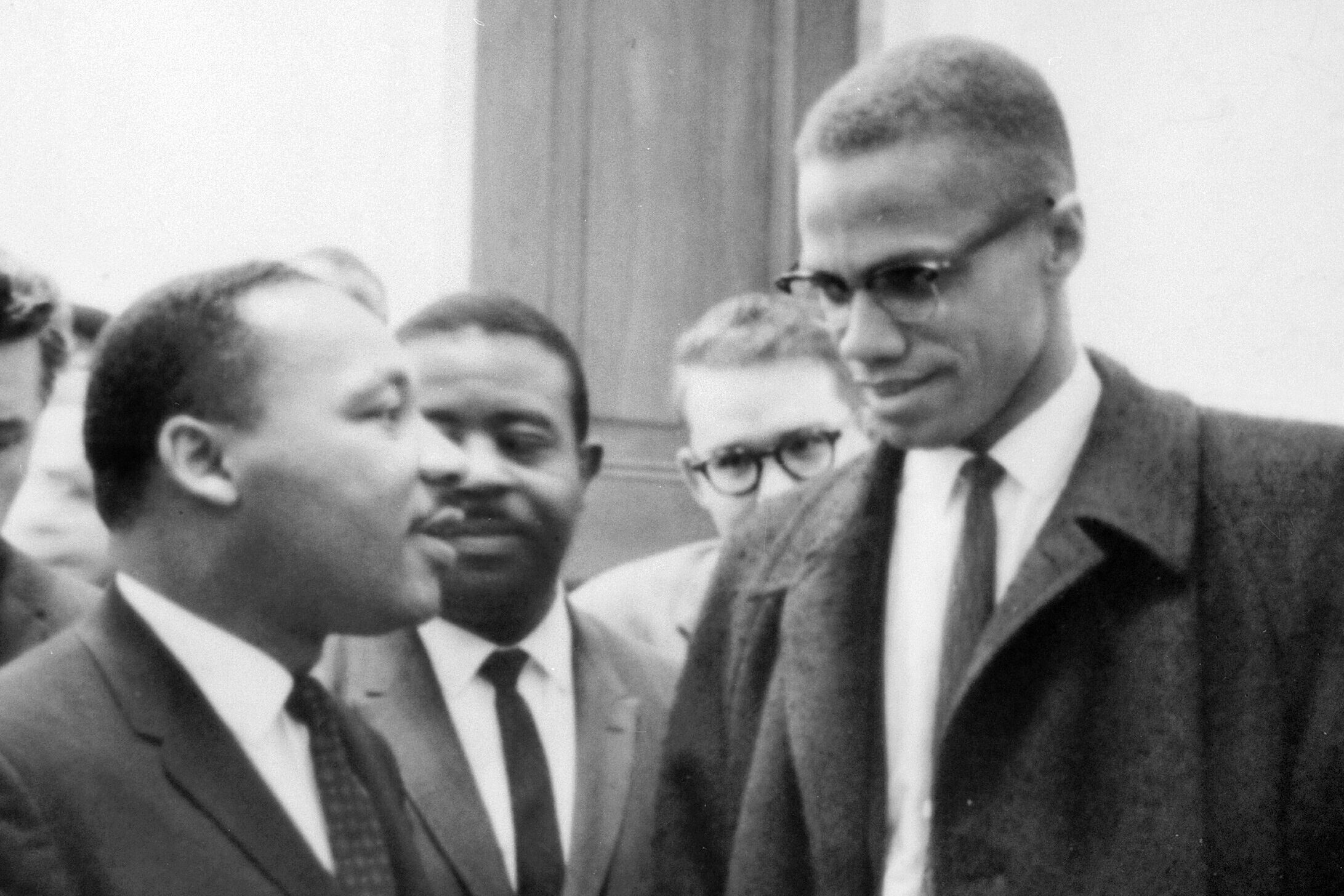 Martin Luther King, Jr. and Malcolm X met before a Senate debate on the Civil Rights Act of 1964. King on the Left wears a dark suit, tie, and a white shirt. To the right, a taller Malcolm X appears in a dark suit and tie, jacket open and a white shirt. Their approach to Civil Rights was very different.
