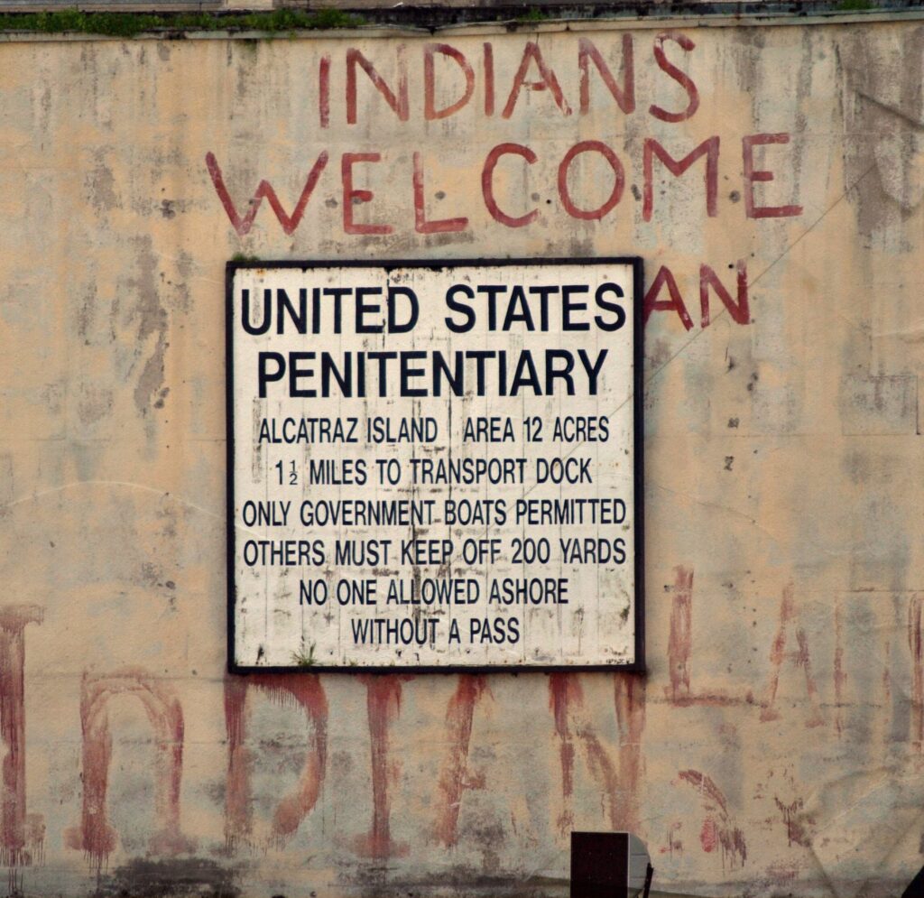 At the center of this image is a sign that reads, "United States Penitentiary. Alcatraz Island. Area 12 acres. 1 1/2 miles to Transport Dock. Only Government Boats Permitted. Others Must Keep Off 200 Yards. No one Allowed Ashore Without A Pass." Behind the sign on the concrete, intertribal members of Indians of All Tribes painted "Indian's Welcome" and "Indian Land" in protest of U.S. land policies that stated abandoned federal land was to be returned to American Indians per the Treaty of Ft. Laramie in 1968.
