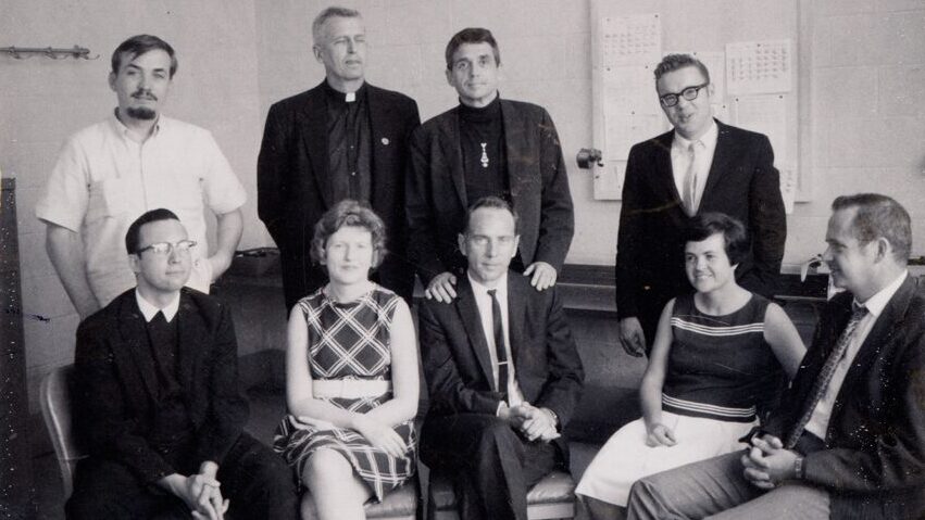 Shown in the black-and-white photograph taken at the Wilkens Police Station are the Catonsville Nine Catholic activists who protested the Vietnam War on May 17, 1968, by burning draft files from a draft office in Catonsville, MD. Pictured from left to right standing, are George Mische, Philip Berrigan, Daniel Berrigan, and Tom Lewis. Left to right, seated are David Darst, Mary Moylan, John Hogan, Marjorie Melville, and Tom Melville.