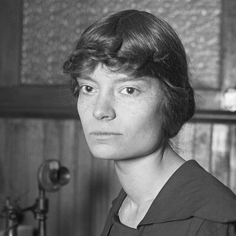 Black and white photograph of Dorothy Day, cofounder of the Catholic Worker Movement to aid the poor and unhoused. She is seated next to a table with a telephone in the background. Day is seen wearing a dark, conservative dress.