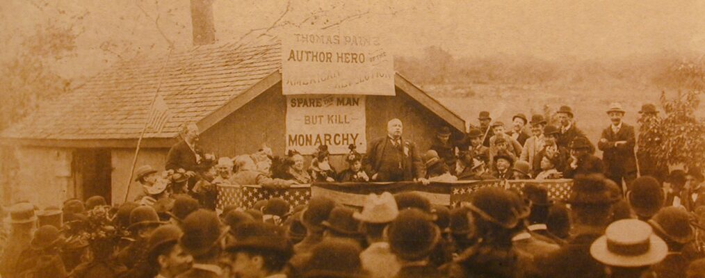 American orator and Illinois Attorney General Robert G. Ingersoll is speaking to an audience in New Rochelle, New York, 1894. The stage is covered with banners of stars and stripes. In the background, two signs affixed to a building read, "Thomas Paine. Author Hero of the American Revolution and below that, "Spare the man but kill monarchy."