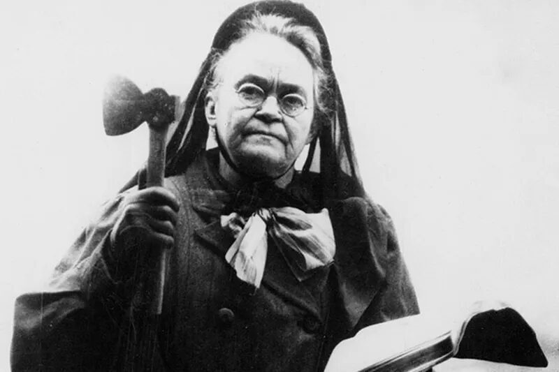 Carry A. Nation with her Bible, open in her left hand, and her hatchet in her right hand, not long before she died in 1911. She is shown in a heavy woolen coat, dress with a bow, and hat trailing down her back.