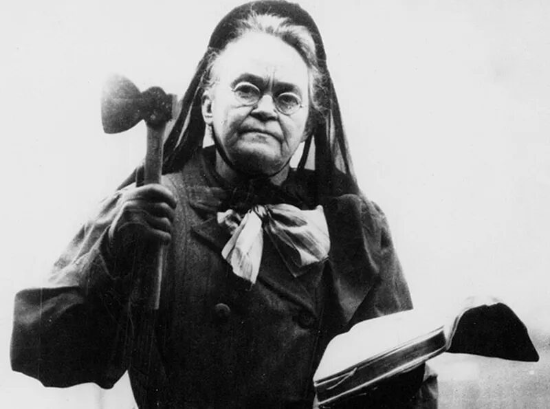 A photograph of Carry A. Nation with her Bible open in her left hand and a raised hatchet in her right. He is wearing a head covering flowing down her back and a dark dress with scarf.