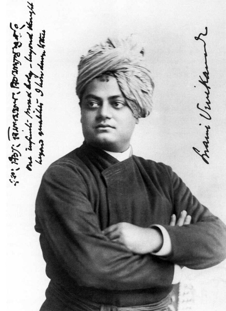 A black-and-white photograph of Swami Vivekananda with arms folded and a turban upon his head. He wears a kurta with a sash around his waist. Photograph taken in 1893 in Chicago. On the left, he has written in what appears to be a phrase in a combination of Hindi and English, "One infinite pure and holy – beyond thought beyond qualities I bow down to thee." Running up the right side is "Swami Vivekanand."