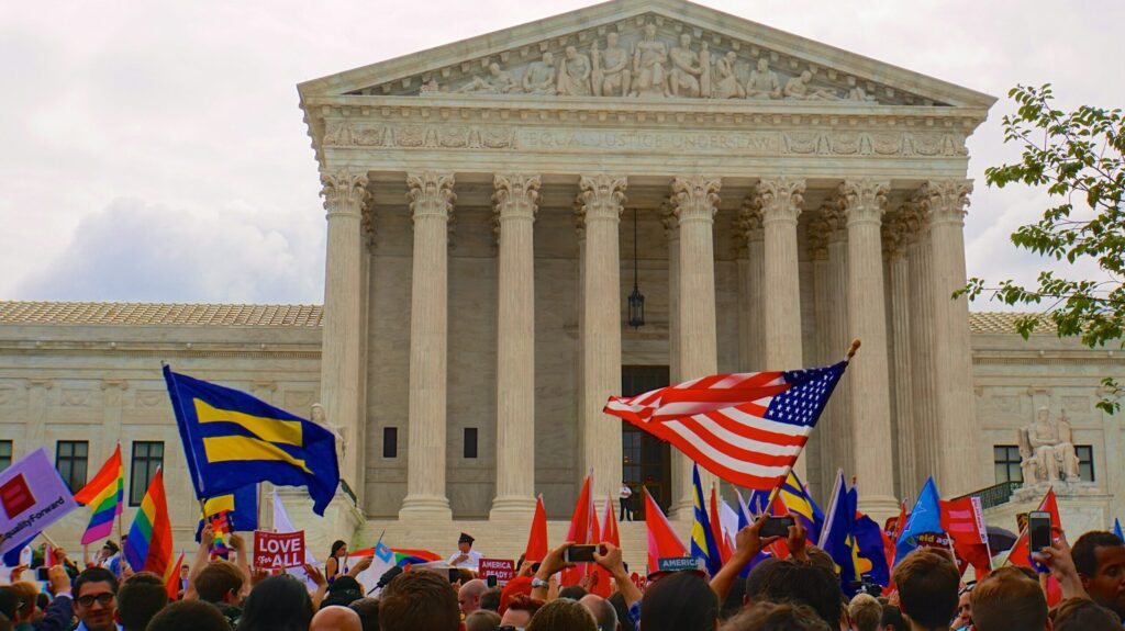 Photograph of a crowd celebrating the Oberfell decision outside the Supreme Court in June 2015. Crowd members hold flags of the United States, a rainbow Pride flag, and a Human Rights Campaign flag that is blue with yellow stripes. The case ended marriage discrimination.
