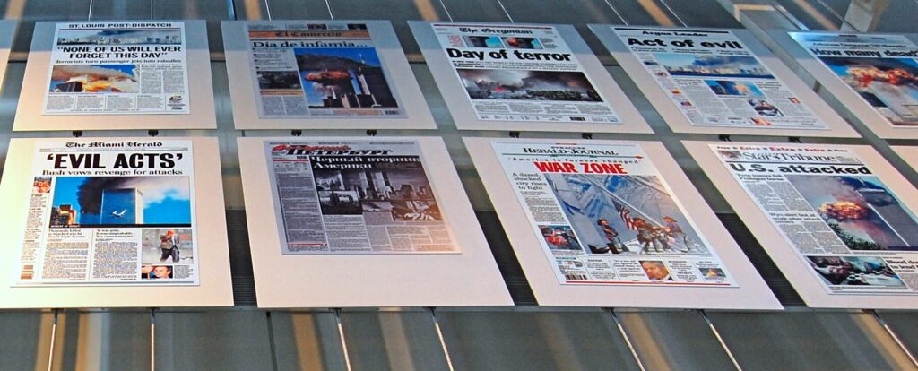 A museum panel depicts newspaper headlines showing reactions to the September 11th attacks. The headlines from the top row, left to right, read:
St. Louis Post Dispatch, "None of Us will Ever Forget this Day." 
Peruvian newspaper "El Comercio,  "Dia de infamia..." 
The Oregonian, "Day of Terror." 
Argus Leader, "Act of Evil."
Arkansas-Democrat Gazette, "How many dead?"
2nd row:
The Miami Herald, "Evil Acts: Bush vows revenge for attacks."
2nd one is in a foreign language
Herald Journal, "America is forever changed. War Zone."
Star Tribune, "U.S. attacked."

