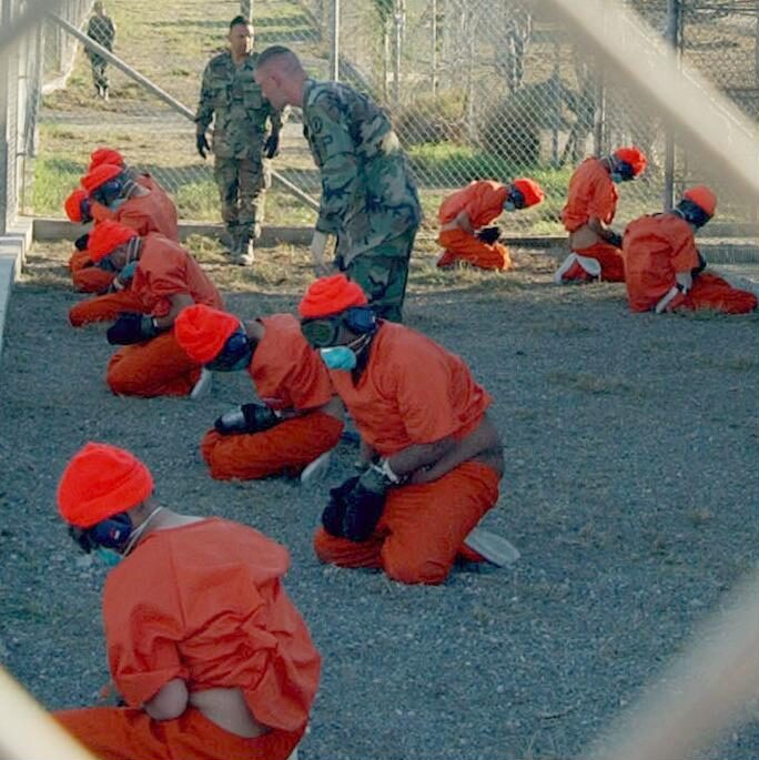 A photograph of ten detainees at Camp X-Ray at the Guantanamo Naval Base in Cuba, 2002. The detainees are cuffed and their eyes and ears covered while squatting on the ground. Each wears orange clothing and hats. Three military personnel are seen inside the fence. One appears to be angrily speaking to the prisoners.