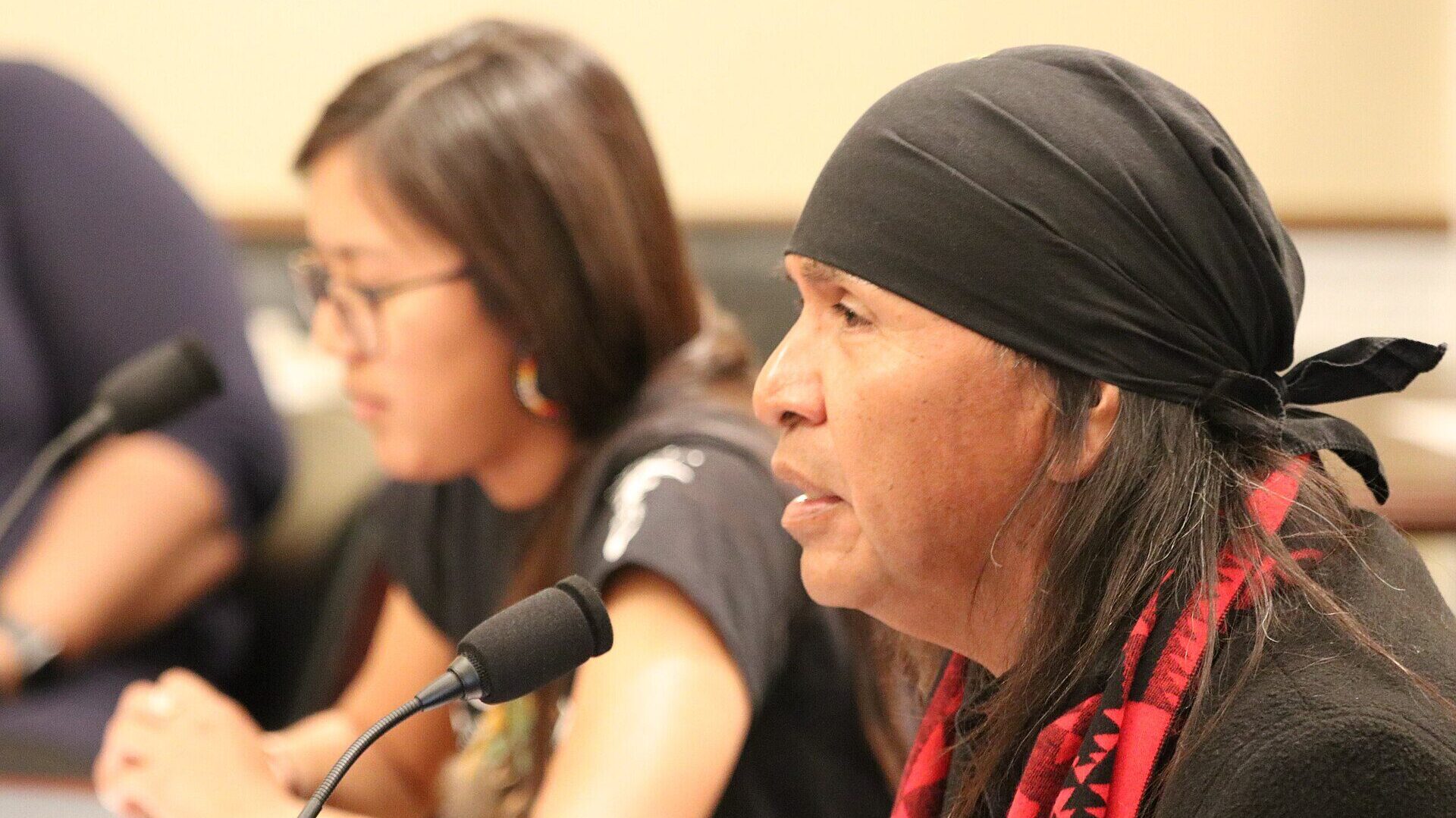 Apache leader Wendsler Nosie Sr. testifies before the United States House Committee on Natural Resources on March 12, 2020, protesting the Oak Flat Mining operations in Arizona. Nosie is pictured at a microphone wearing a black coat, a red and black scarf with a geometric imprint, and a black scarf on his head.