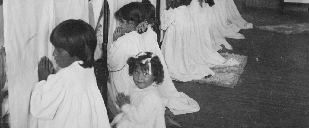 This photograph depicts young children, likely Mojavi or Hopi, at the Phoenix Indian School in Arizona. The girls are kneeling before their beds with folded hands, saying prayers. 