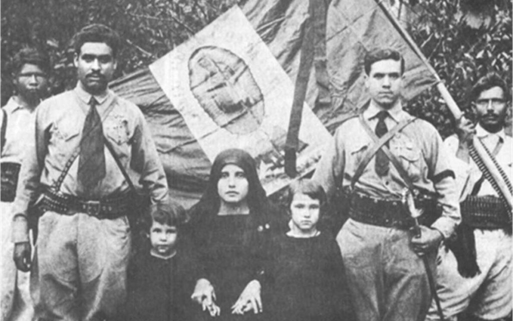 This photograph shows three officers in a Cristero regiment in full uniform standing with their families. Small children are in the foreground center. The flag in the center background depicts the Virgin of Guadalupe. The Cristero War mobilized Catholics in the United States to support the Church in Mexico, and it also brought an influx of refugees to the American Southwest, further entrenching the Mexican Catholic presence in the region.