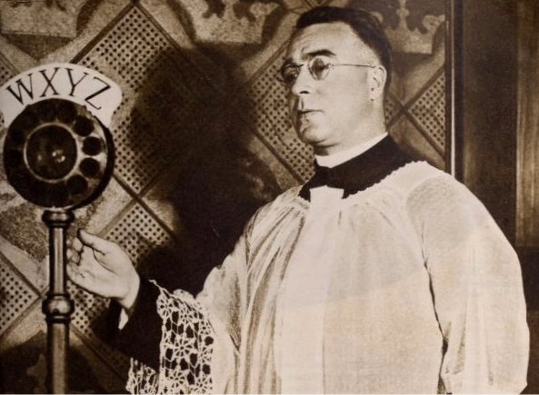 Photograph of Father Coughlin, a Catholic priest and radio personality, delivering his Golden Hour program, 1934. Coughlin is seen wearing a priest's frock covered with a white gown with intricate lace. His outstretched hand is near the radio microphone.