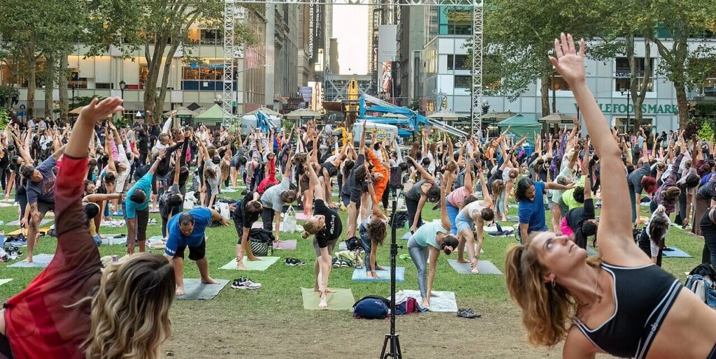 This photograph shows Americans practicing yoga at Bryant Park, New York, 2023. In the background stand tall office buildings and storefronts. To the left is the "Whole Foods Market."