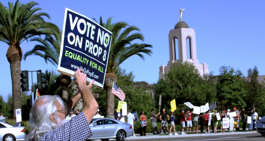 A protester with a checked shirt and long white hair holds an anti-Proposition 8 sign in the front left corner of the photograph. Across the road is a line of protesters also holding signs, but they are unreadable.