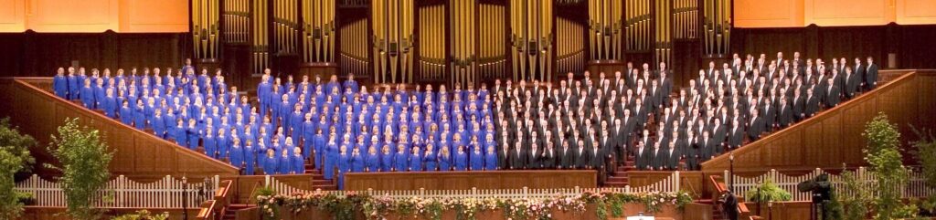 A photograph of the Mormon Tabernacle Choir in Salt Lake City. Photograph taken in 2004. Women wear royal blue dresses and men black suits. Beautiful pipe organs fill the center of the image. 