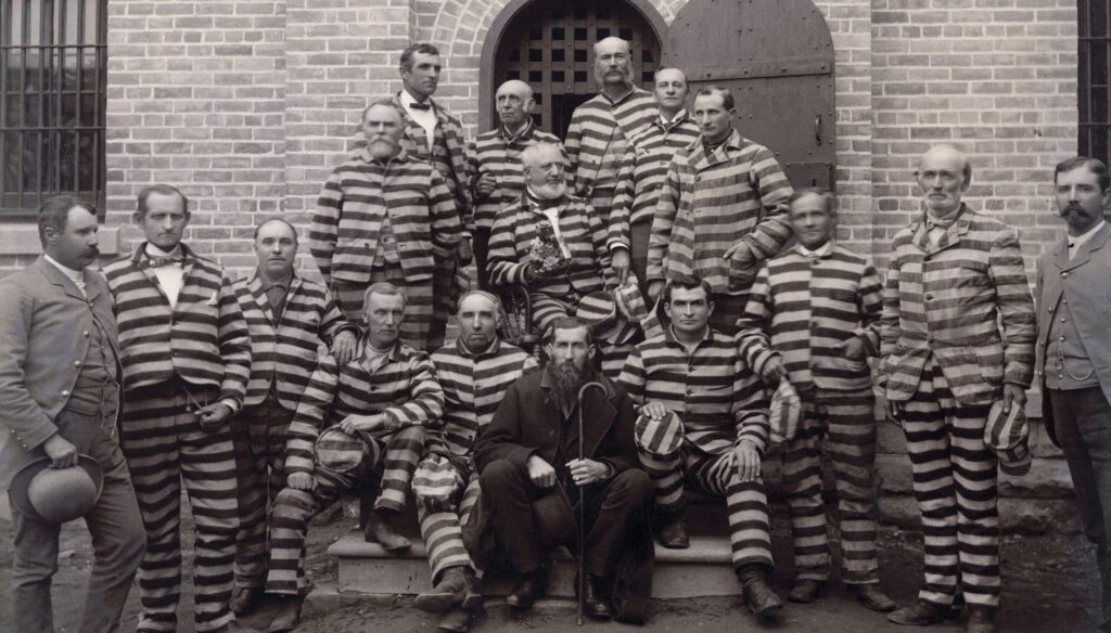 A group of Mormon polygamists dressed in striped clothing is seen in a prison in this photograph by Charles Roscoe Savage, 1889. One prisoner holds a bouquet. Three gentlemen are seen in suits, one to each side and one in the center holding a cane.