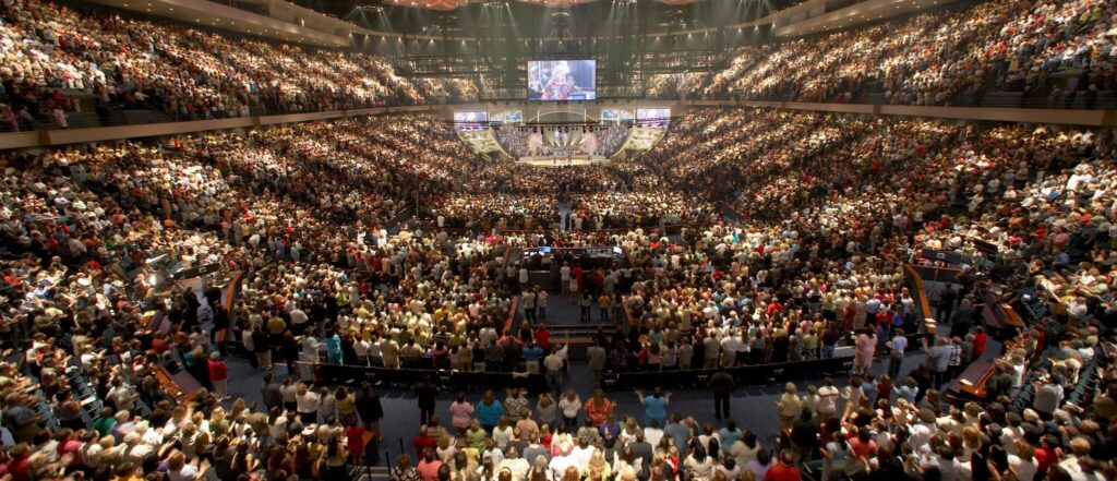 This photograph of Lakewood Church in Houston, Texas illustrates the massive popularity of the prosperity gospel. The building is filled with hundreds of people standing. A stage and television are in the upper center of the photograph.