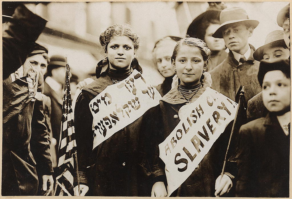 A black-and-white photograph of children at a labor parade in New York City, about 1909. Two girls wear banners with "Abolish Ch[ild] Slavery!" in Yiddish and English. One girl carries an American flag and the other a black flag.