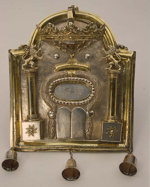 An ornamental Gilt silver Torah breastplate from 1810 is depicted in the photograph. Hanging from the bottom are three bells.