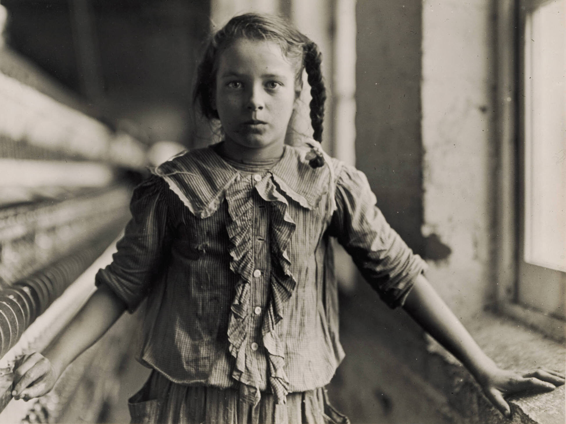 A young girl can be seen in this photograph with a pigtail to one side and a ruffled blouse, standing with her hands on a wall and spinning machine. She works in a Carolina Cotton Mill, 1908.