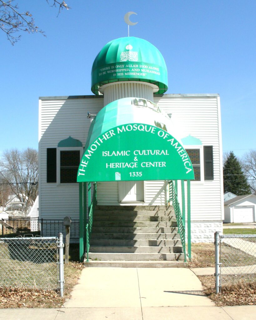 A small white structure is identified as "The Mother Mosque of America. Islamic & Heritage Center. 1335" on the green awning over concrete steps that lead into the main building. The green dome on the top of the main part of the mosque is topped with a crescent moon in silver and has the phrase, There is only Allah (God Alone) to be worshipped, and Muhammed is his messenger." 