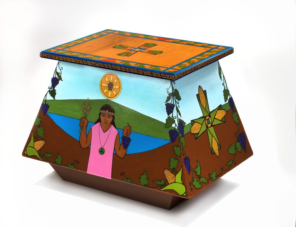 This "Farm Worker's Altar" (1967), by Emanuel Martines, is hand-painted in a vibrant, folk style. It has a rectangular lid with a yellow background, a blue geometric border, and a multicolored cross at the center. The lower part of the box is sloping downwards and features a brown-skinned Christ and, on another, an indigenous woman wearing a green and orange headband, a peace symbol necklace, and a pink tunic. In one hand she holds a bunch of purple grapes and in the other small wheat stalks. AT the top is a symbol of the sun, and on the border is a grapevine and a couple of ears of corn on each bottom corner. On the side panel, a cross made of four corn ears in their husk forms a cross. This altar was created to commemorate Cesar Chavez's fasts protesting unfair employment practices and unsuitable work conditions.