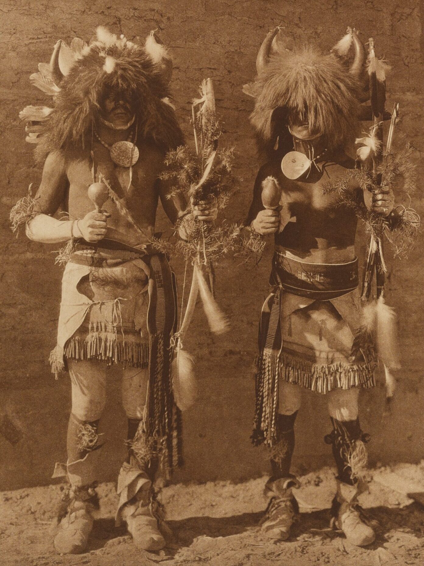 This photograph from Edward S. Curtis shows two Pueblo men dressed to perform a Buffalo dance in 1925, Tesuque, New Mexico. The men wear headdresses of feathers, hair, and horns. Around their necks, they have necklaces with large round medallions, and in their right hands they carry rattles. In their left hands, they hold arrows with dried plants and a feather in a bundle. Around their waists are skirts with fringe, and on their feet they are shown wearing moccasins.
