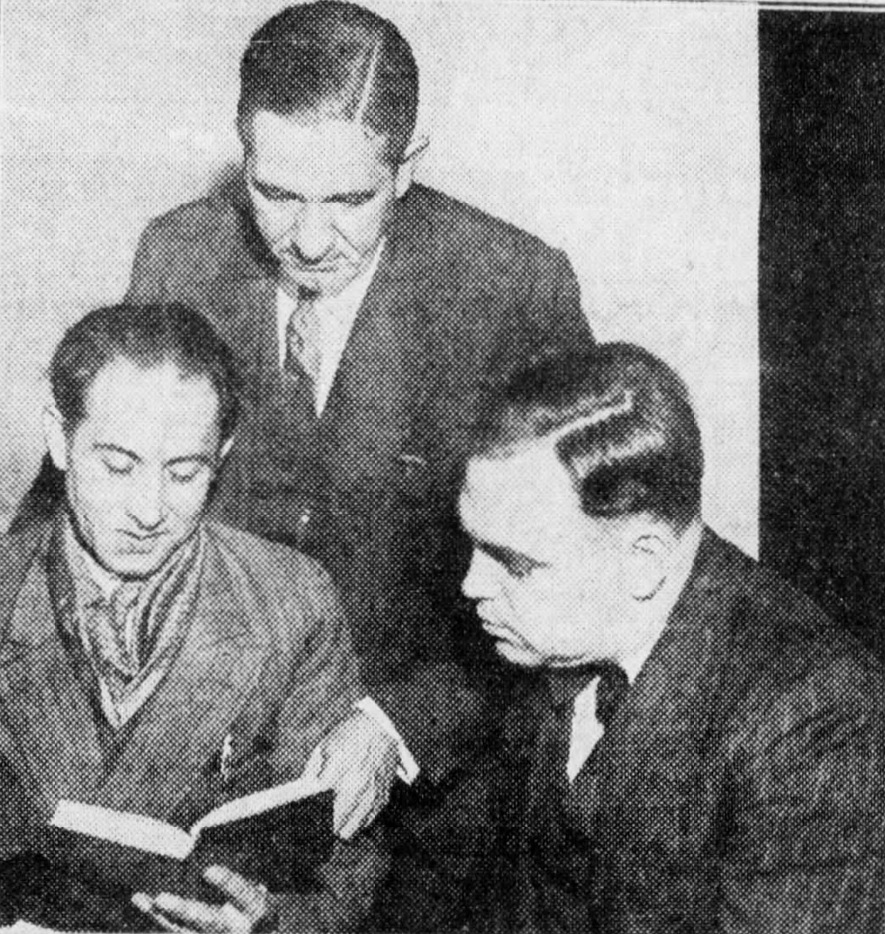 Wallace Fard Muhammad, left, shows a book to police detectives who lean in to see. One detective is seated to his left, and the other is behind Fard, leaning over his shoulder.