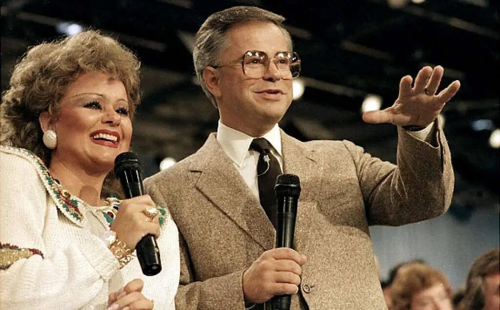 A smiling woman and man stand side by side onstage, each holding a microphone. The woman wears a white outfit with colorful trim and large earrings; the man wears glasses, a tan suit, white shirt, and tie and gestures with one hand. An audience is visible in the background.
