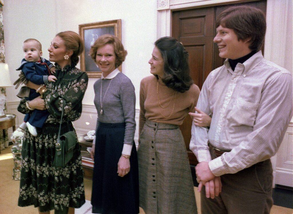 A woman in a green dress, the Shahbanou of Iran, holds a baby in a sailor outfit. Rosalyn Carter looks at the camera while Caron and Chip Carter, the parents of the baby smile and look at their child. 