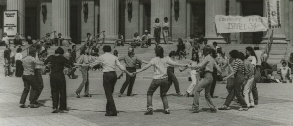 In 1982, Jewish students at the University of Minnesota danced in a circle to celebrate the 34th anniversary of the creation of the state of Israel. In the background, people sit on stairs outside the Northrop Auditorium. In the top right corner of the image is a Jewish flag and the words "Celebrate peace. Israel 34."
